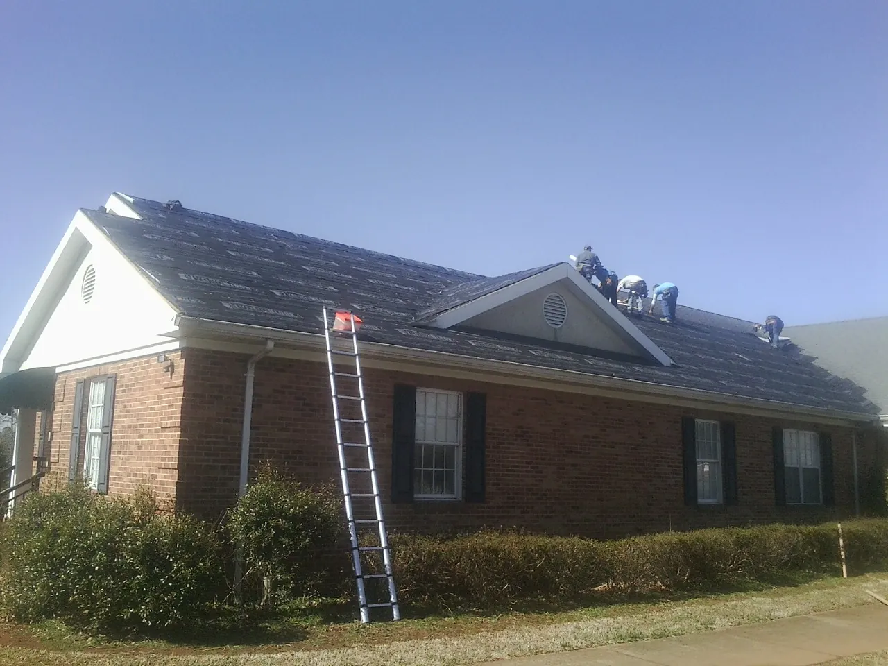 Crew tearing shingles off a brick ranch roof with a ladder set — Emergency Roof Repair work by our crew in Ursa