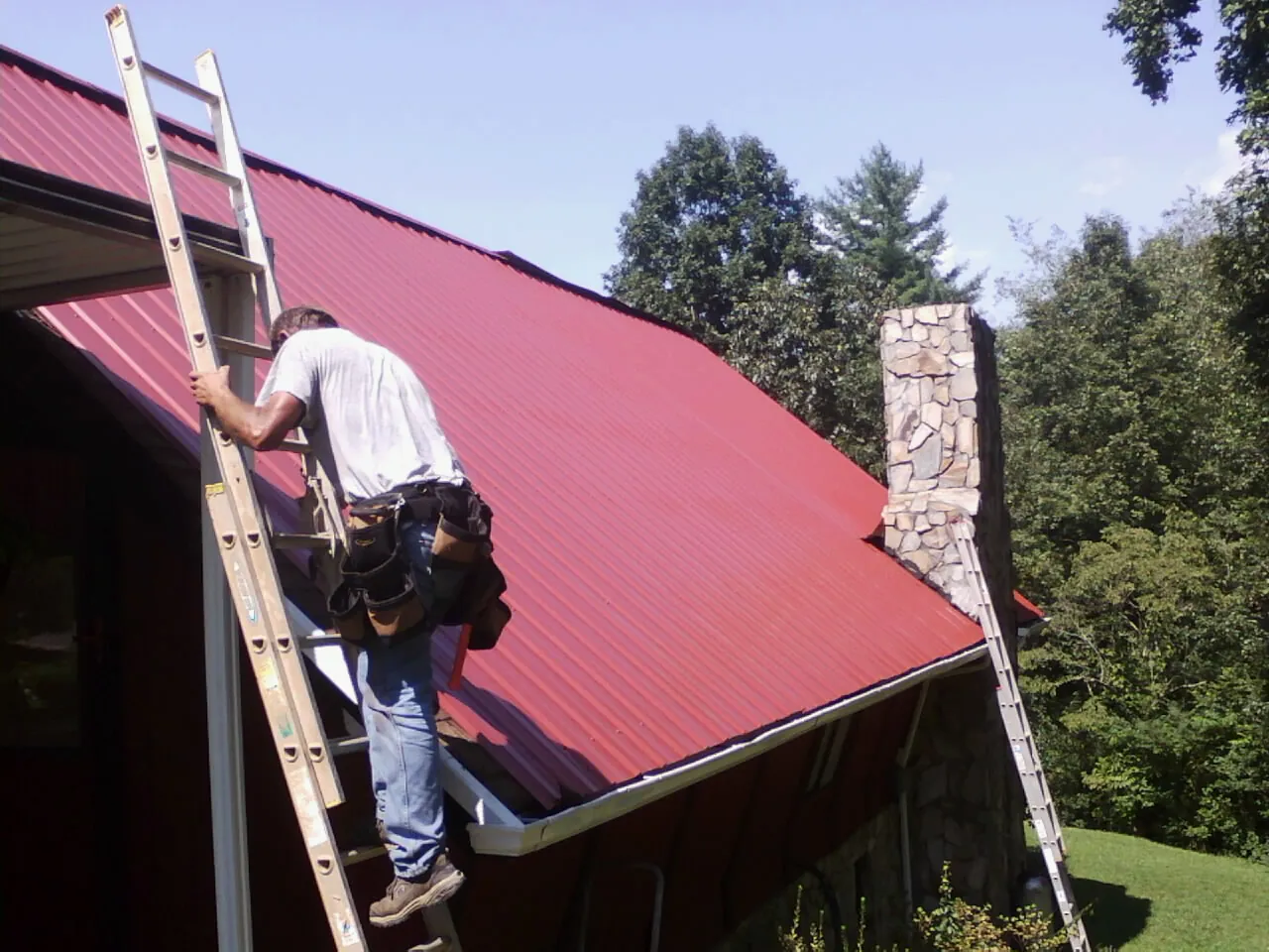 Roofer on a ladder beside a red metal roof and stone chimney — Soffit & Fascia Repair by Roofing Brothers in Ursa